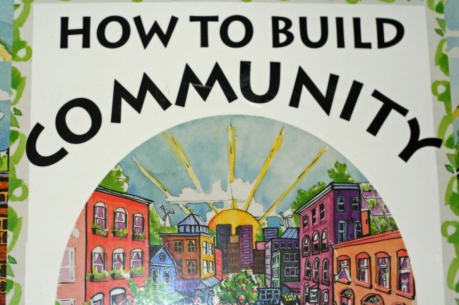 A photo of the front cover of a book called How To Build Community with a picture of a hand drawn cartoon town, made up of buildings with lots of colors, trees and rooftop gardens around them, and a bright sun rising over the horizon.