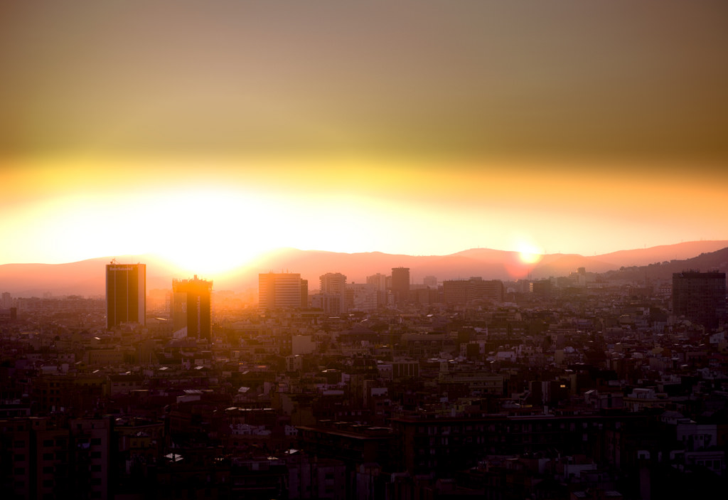 An edited photo of a heavily industrialized city with many skyscrapers, and a sensation of a desert in the distance and lots of air pollution, all stemming from what it likely an explosion of some kind far away that is large and bright, like a setting sun