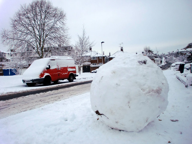 A photo of a giant snowball sitting on the side of a road; background shows a red truck covered in show, a large tree, and houses far off. Everything is covered in snow.
