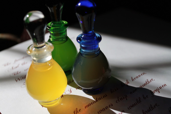 a photo of 3 potion bottles arranged on top of a paper with calligraphy on it, casting deep shadows across the foreground. The closest bottle contains foggy pink liquid, and the other two bottles are blue and green.