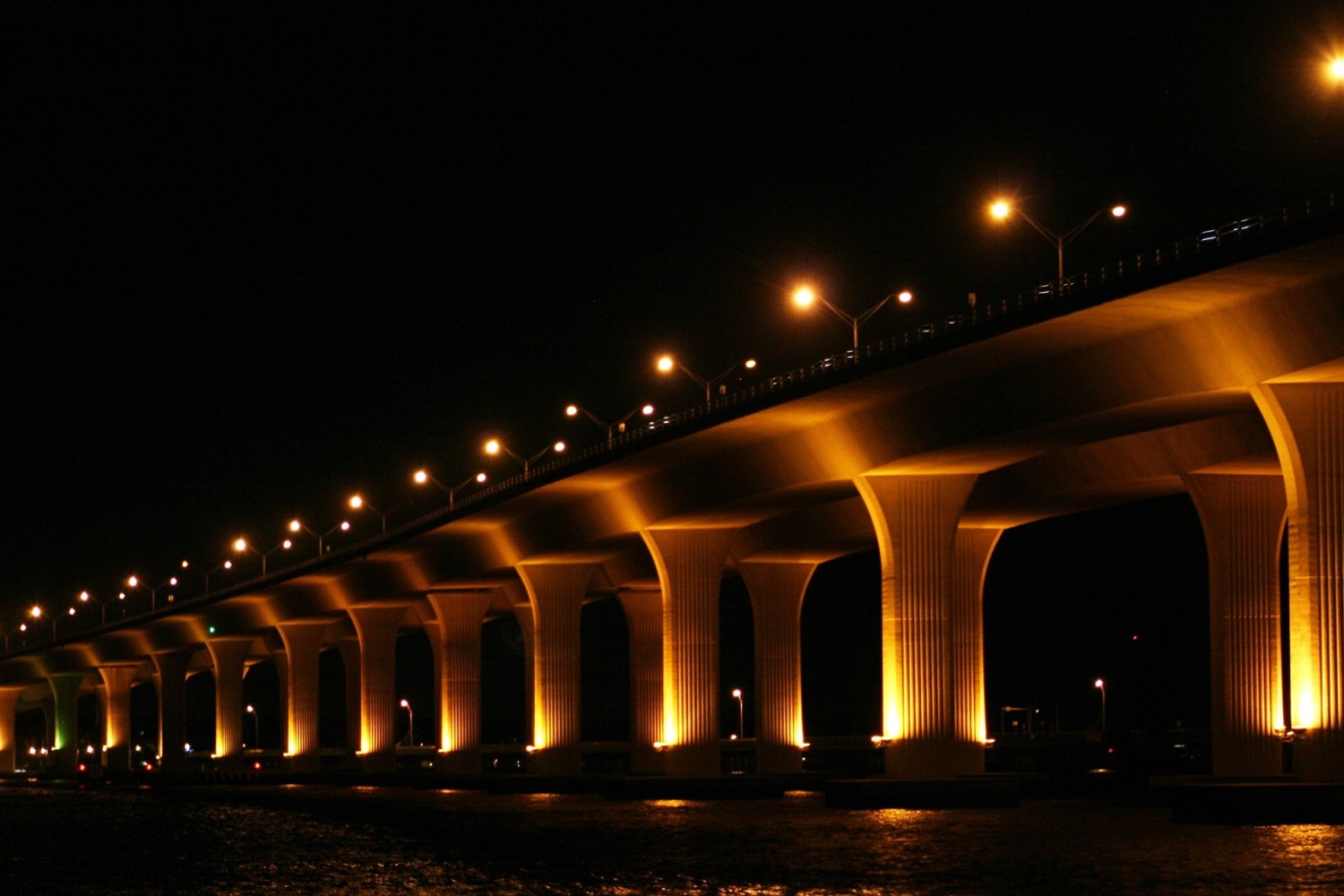 Photo of a bridge lit with yellow lights, focused on the stone supports underneath. At night.