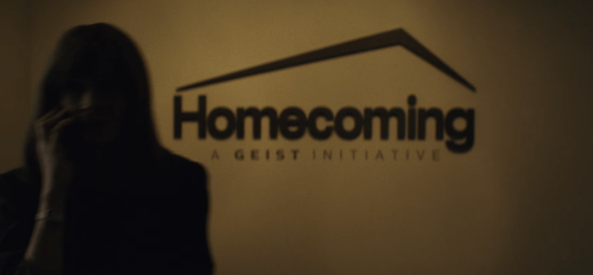 screenshot of a woman on a phone in a blurry foreground; in the background a wall with a sign that says HOMECOMING, A Geist Initiative