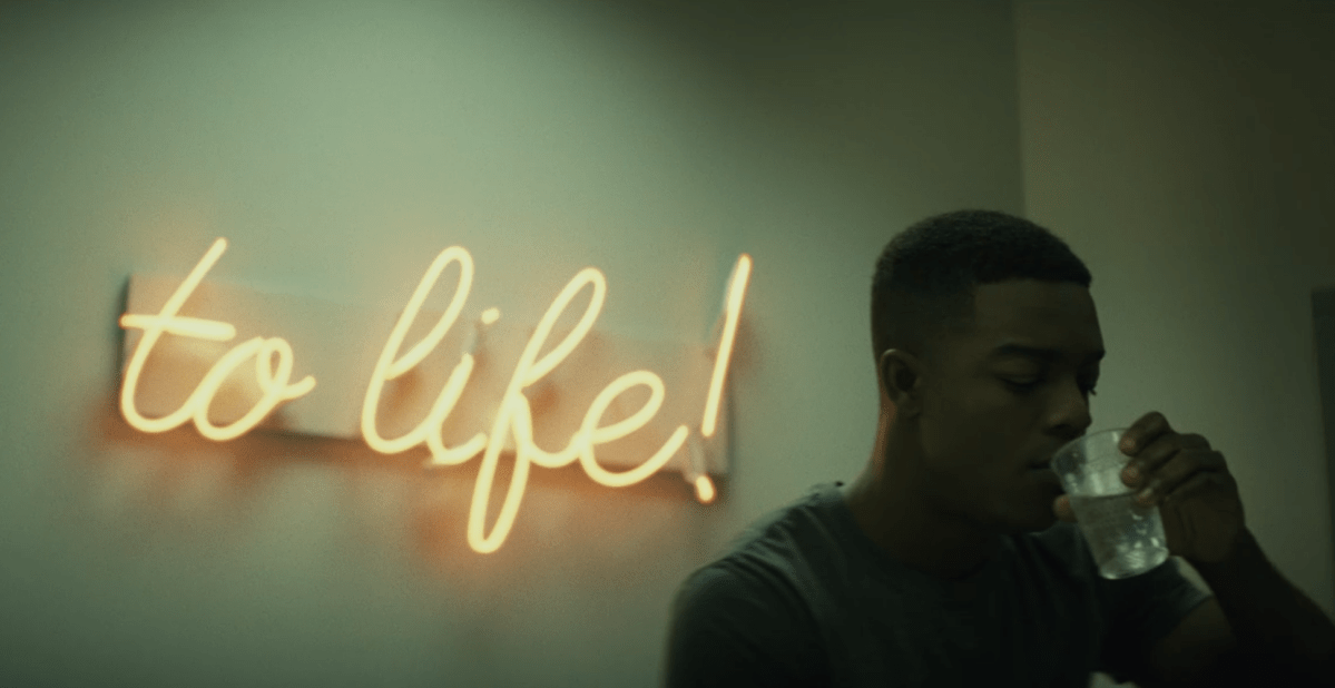 screenshot of a neon sign that says "to life!" in cursive with a young man drinking water next to it.