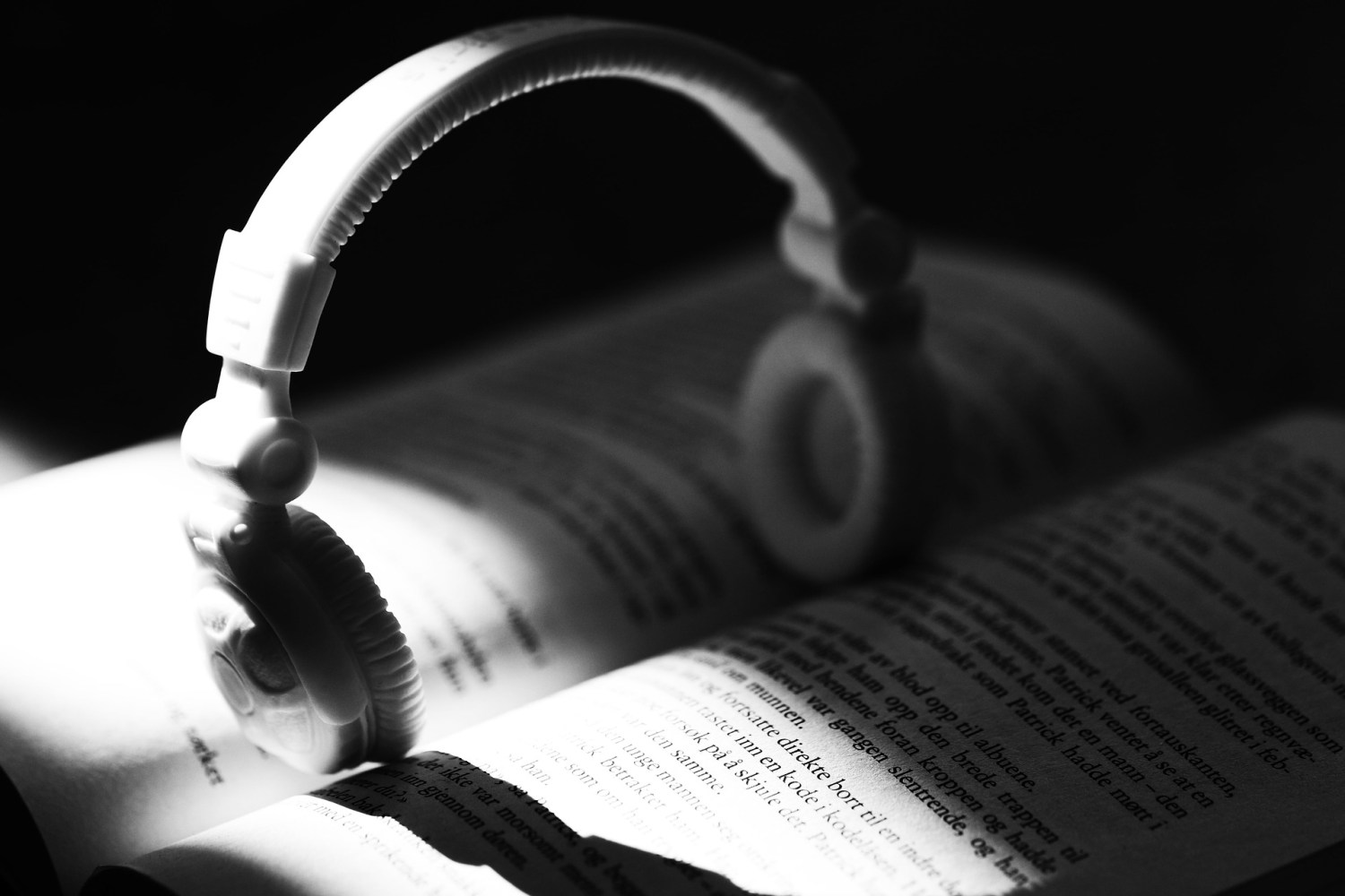 black and white photo of a pair of white headphones standing upright in the open spine of a book, sunlight cast across the bottom half.