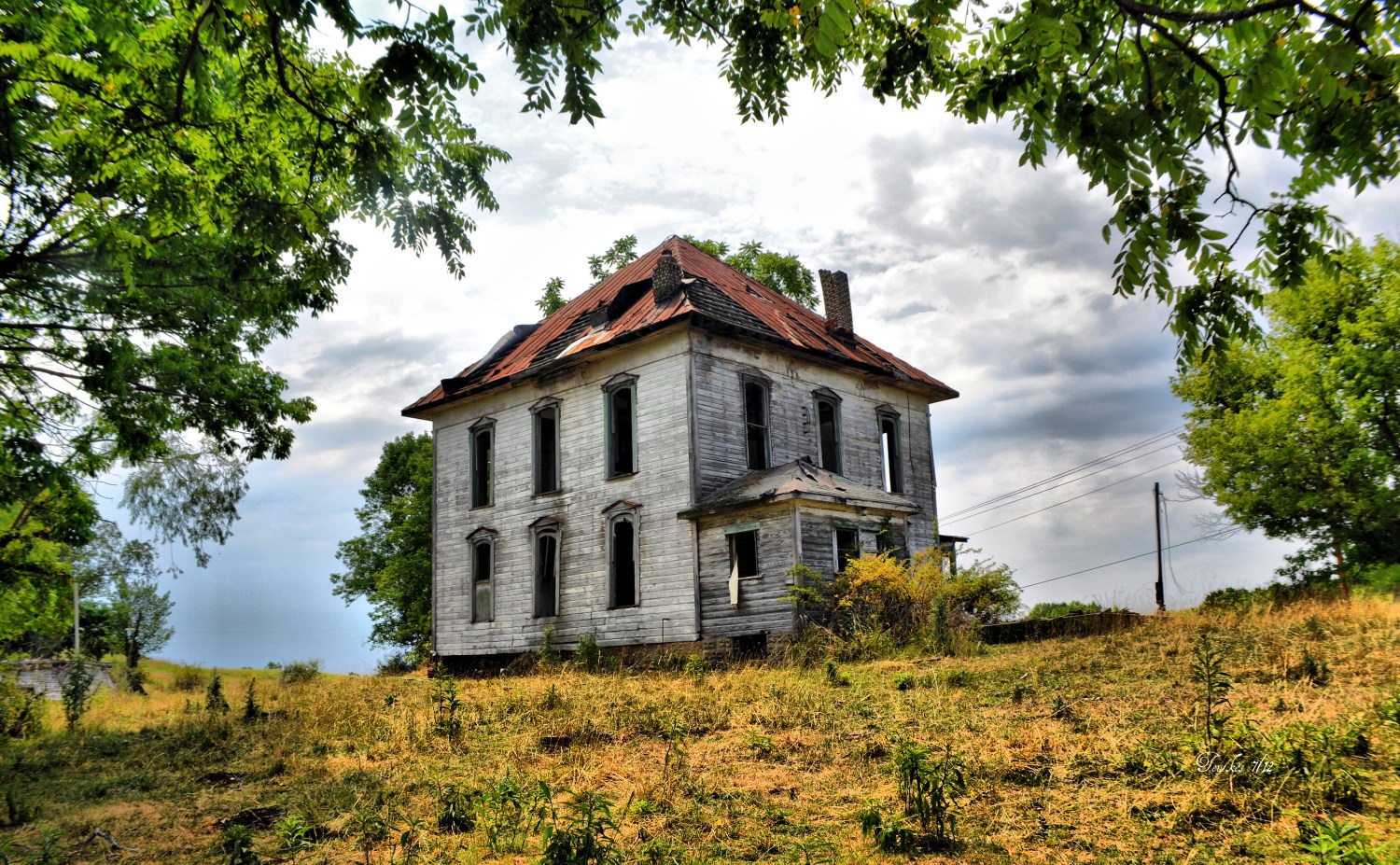 a decaying tall hous, three story wood house in the middle of nowhere, with a greying sky, but brightly colored surroundings, creating a sensation of naivety against the rampant onset of decay.