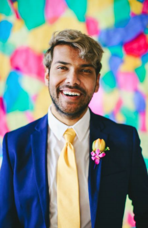 headshot of Tau Zaman creator of CARAVAN. They are wearing a nice dark blue suit with a contrasting pastel yellow tie, and a matching yellow flower in their buttonhole, against a colorful background.
