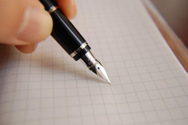a black fountain pen, close-up of the sharp silver tip, held by a white hand, against a blank sheet of graph paper.