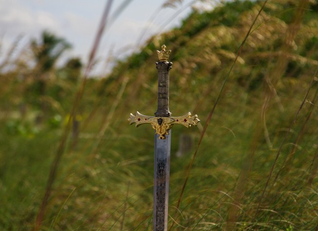a sword with a golden gilt handle abandoned, sticking straight up out of a blurry grassy field.