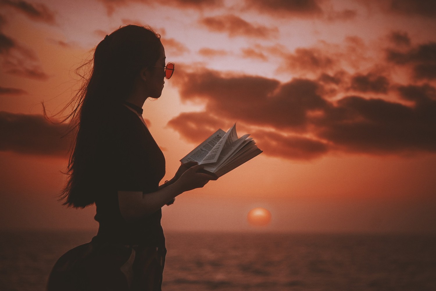 woman wearing sunglasses and reading a book in front of an ocean at sunset